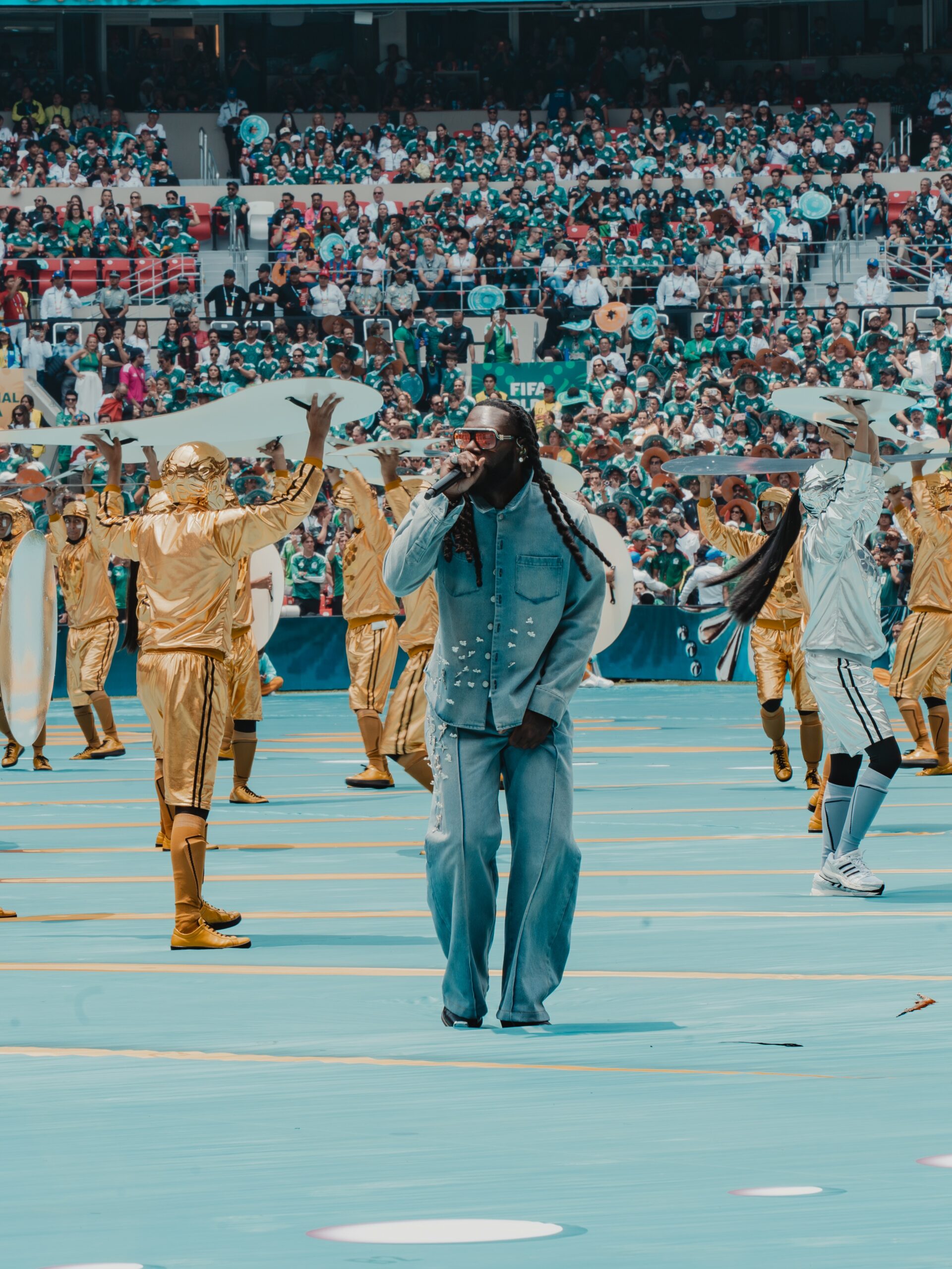 Burna Boy in a blue denim outfit sings into a microphone on a blue pitch, surrounded by dancers in metallic gold and silver costumes with stadium crowds in the background.