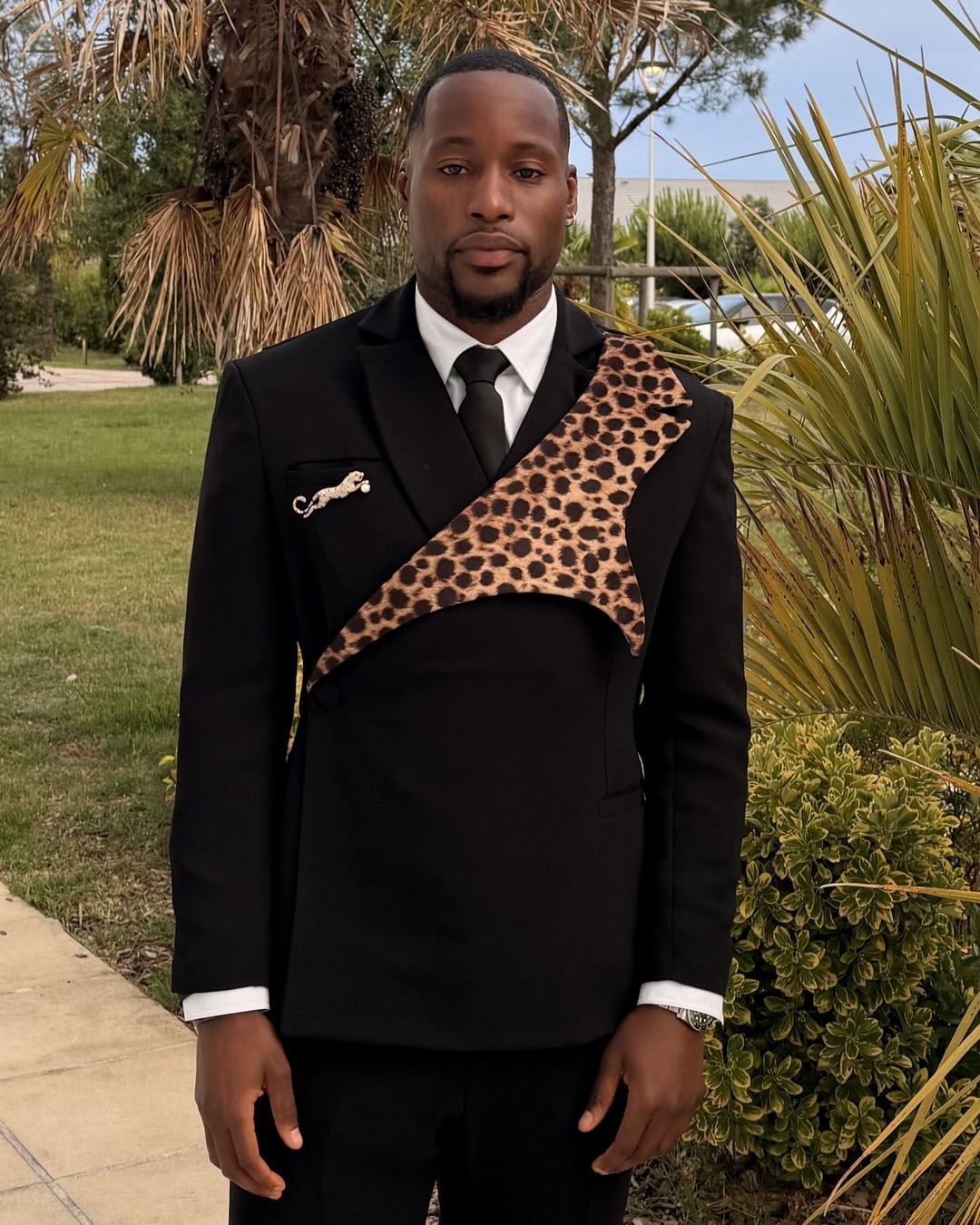 DR Congo goalkeeper Lionnel Mpasi in the official squad arrival outfit featuring a black single-breasted suit with an integrated asymmetric leopard-print panel across the chest and a gold leaping leopard lapel pin.