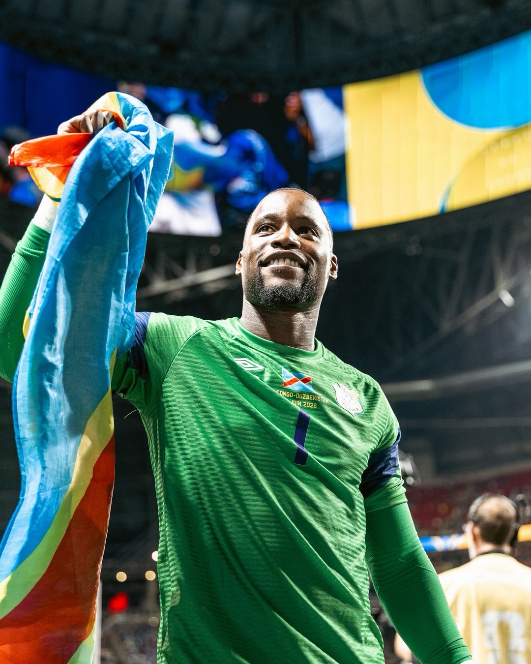 DR Congo goalkeeper Lionnel Mpasi smiling and holding up the Democratic Republic of the Congo flag inside a stadium.
