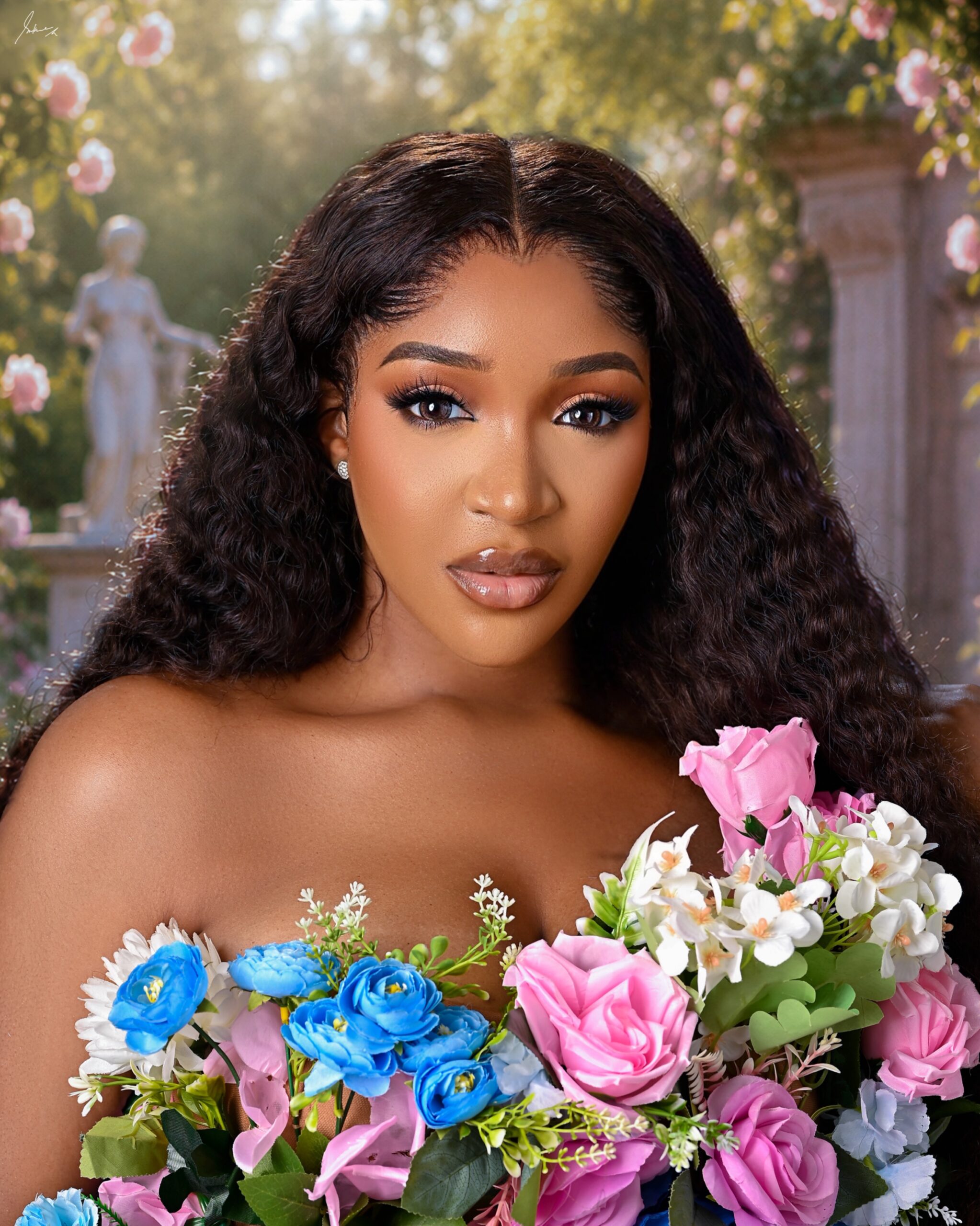 Idia Aisien featuring neutral brown soft-glam makeup and long water-wave curls, wearing a strapless bodice decorated with pink and blue roses.