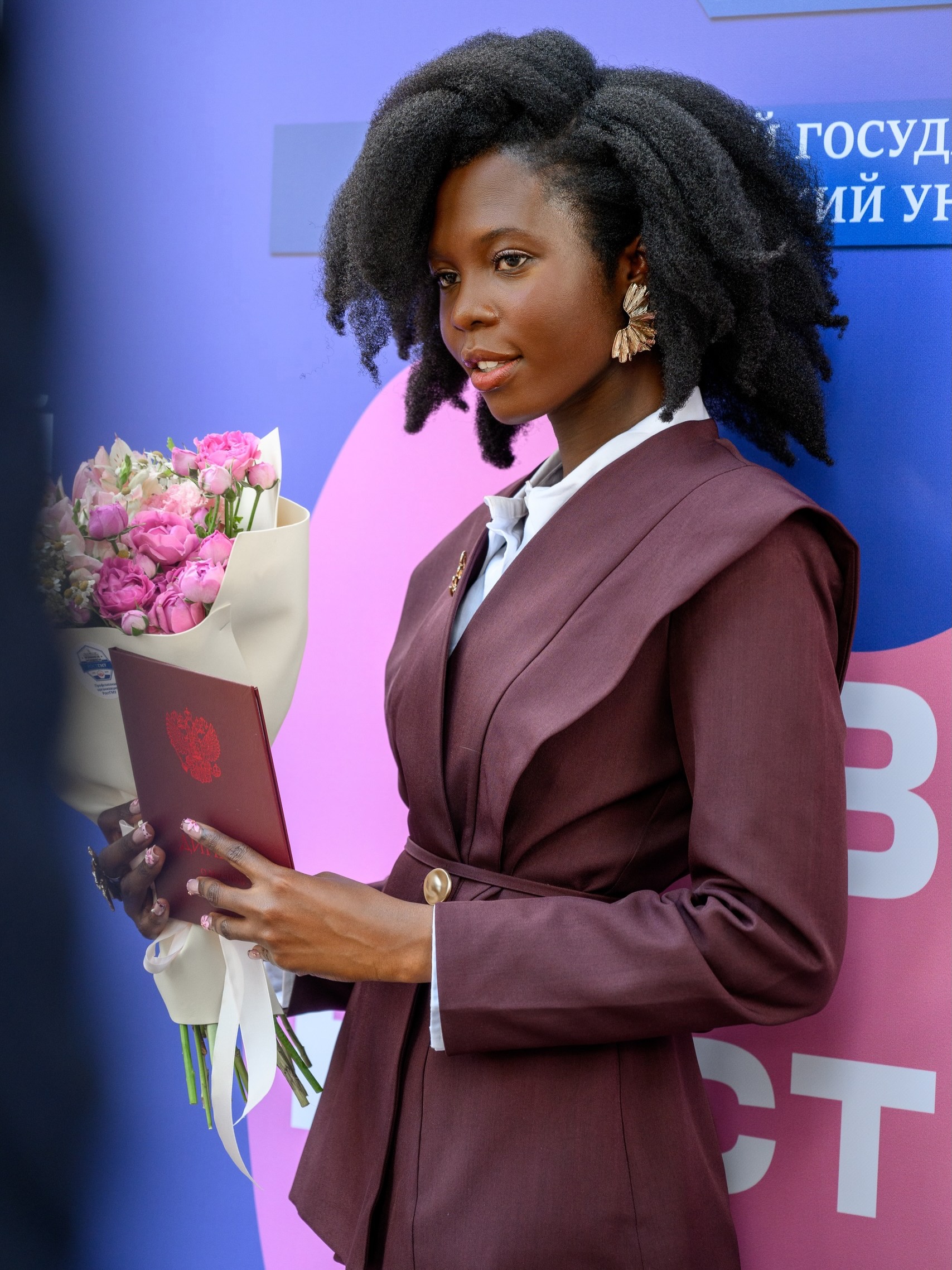 Dr Dorcas Akanbi looks to the side holding flowers and her red medical diploma booklet against a purple background.