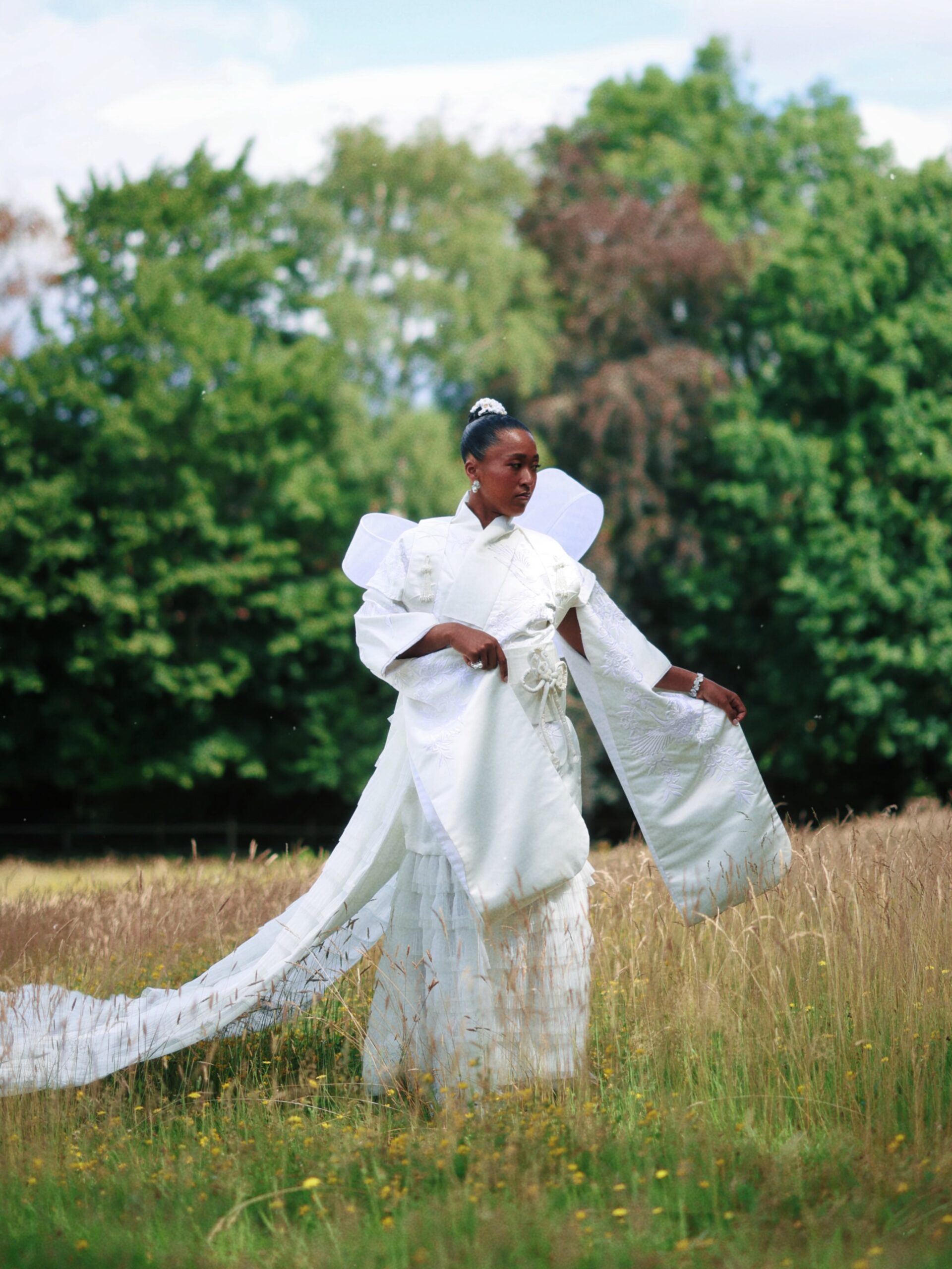 Naomi Osaka posing in a field wearing a white upcycled shiromuku bridal dress gown with a long tulle train.
