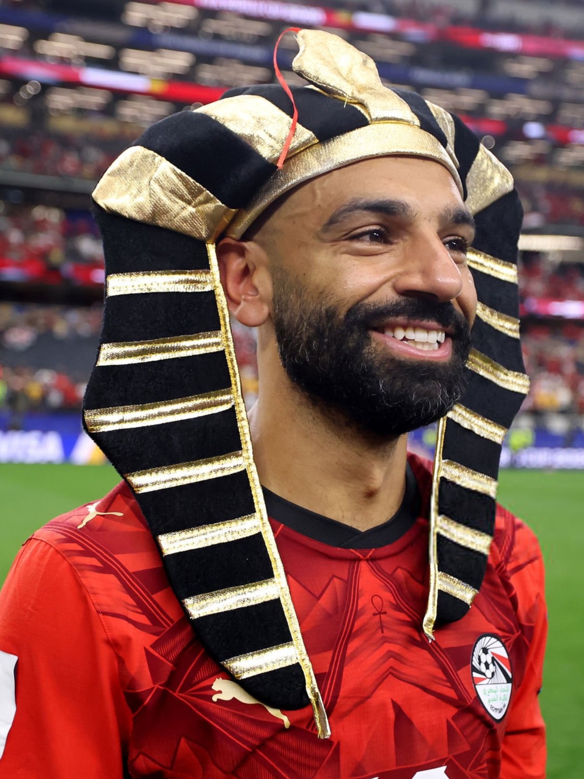 Mohamed Salah smiling on the football field while wearing a black and gold Egyptian pharaoh headdress.