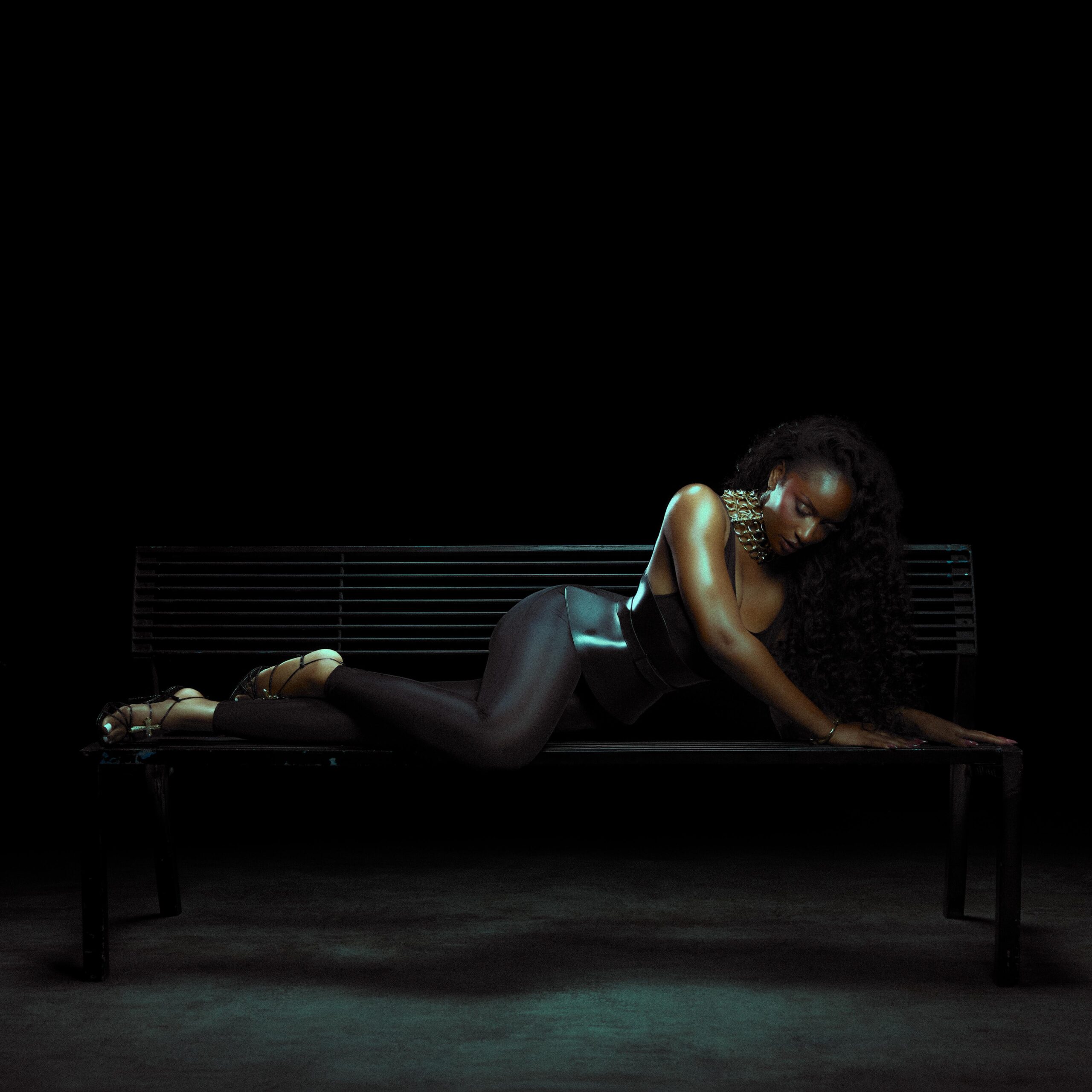 Ayra Starr reclining on a dark slatted bench in a black bodysuit, structural leather corset belt, and second-skin leggings.