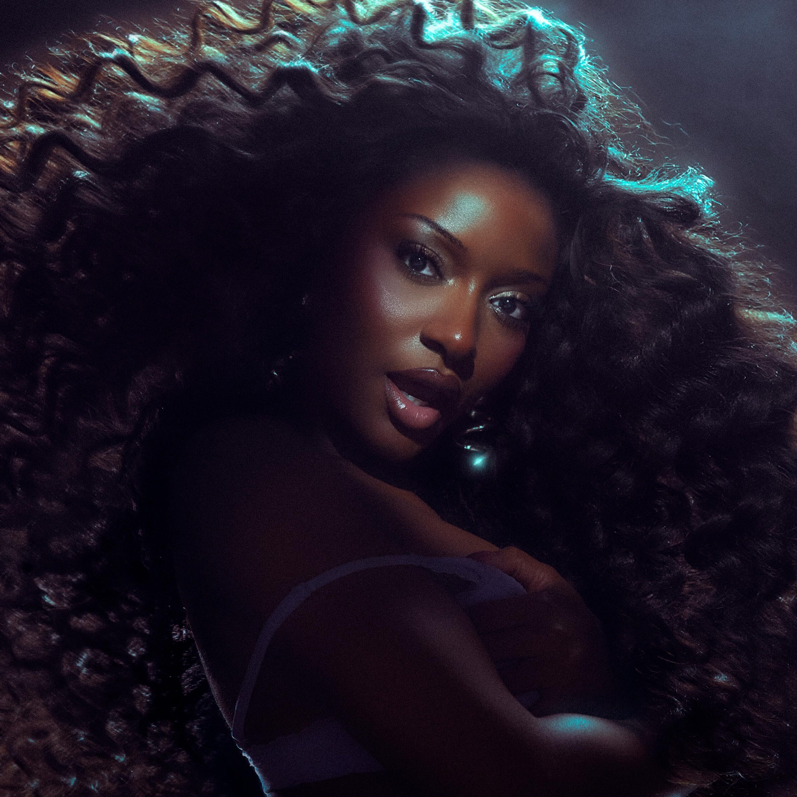Ayra Starr with long cascading curls and a smoky winged eye under moody blue-green studio lighting.