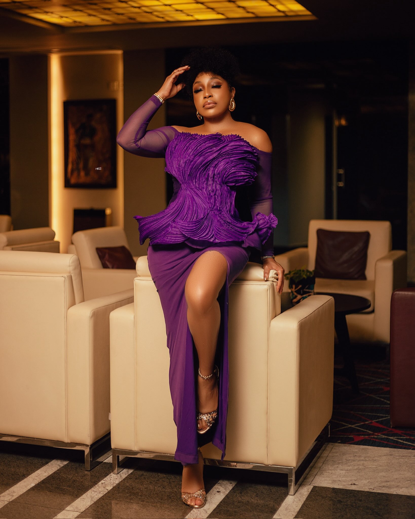 Rita Dominic posing against a leather armchair in an off-the-shoulder royal purple gown featuring a heavily pleated sculptural peplum bodice and a high thigh-slit.