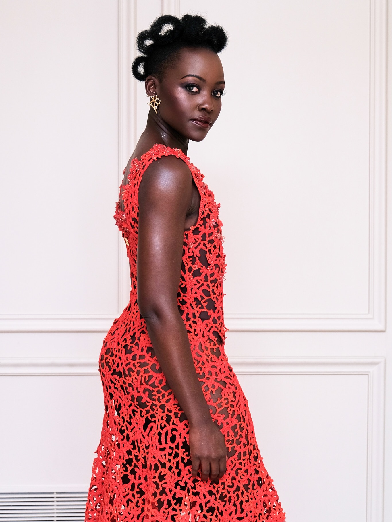 Lupita Nyong'o displaying the side structure of her coral-red Chanel open-work lace gown.