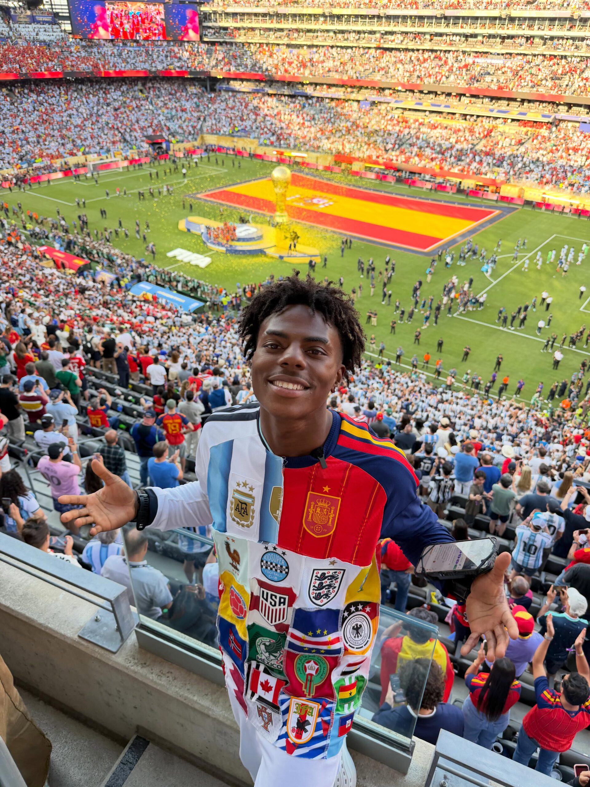 IShowSpeed smiling in the stands at MetLife Stadium wearing a multi-team national football jersey.