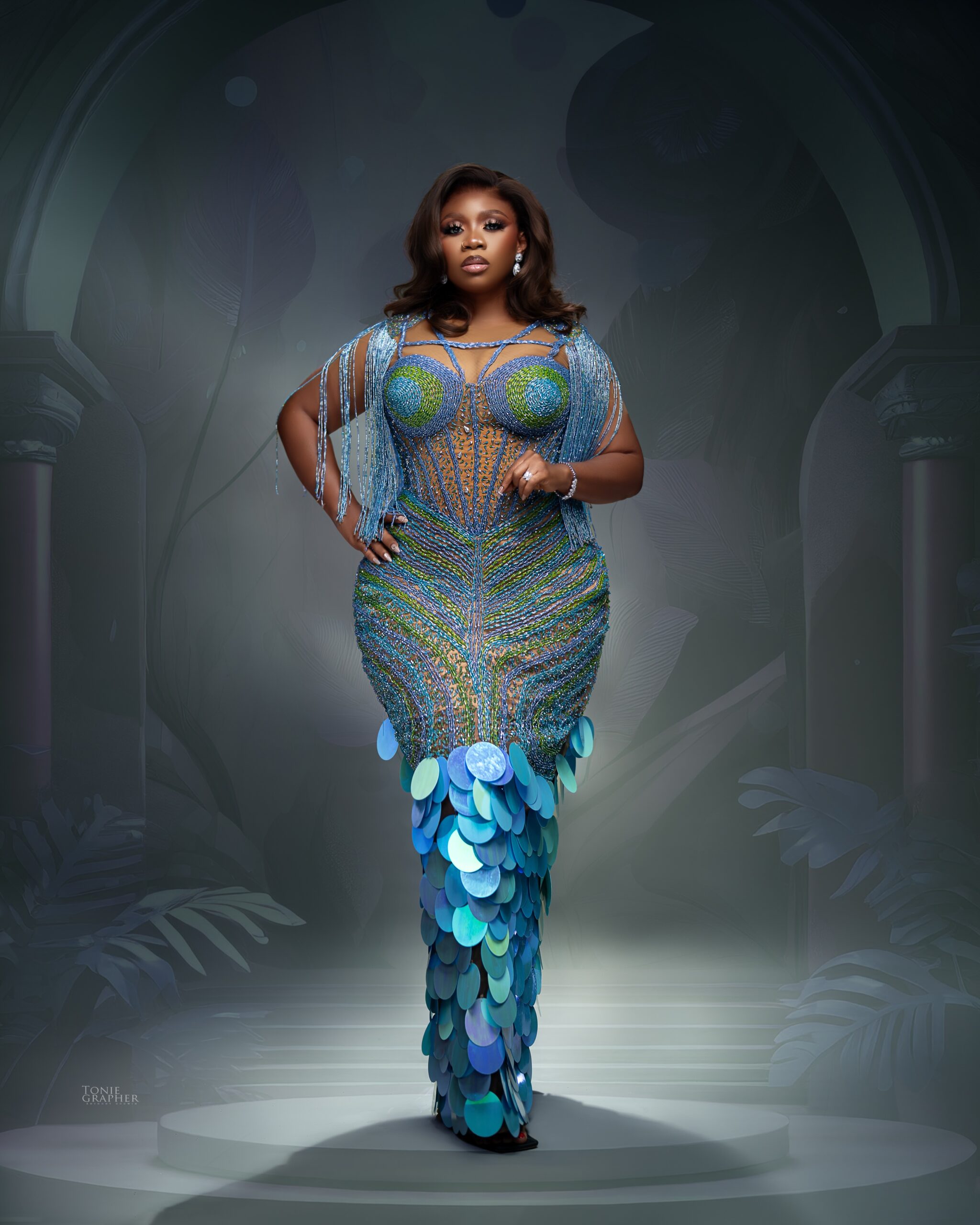 Wumi Toriola wearing a floor-length illusion gown hand-beaded with teal, emerald green, and royal blue wave patterns, featuring crystal fringe capes and iridescent scale-like sequin discs.