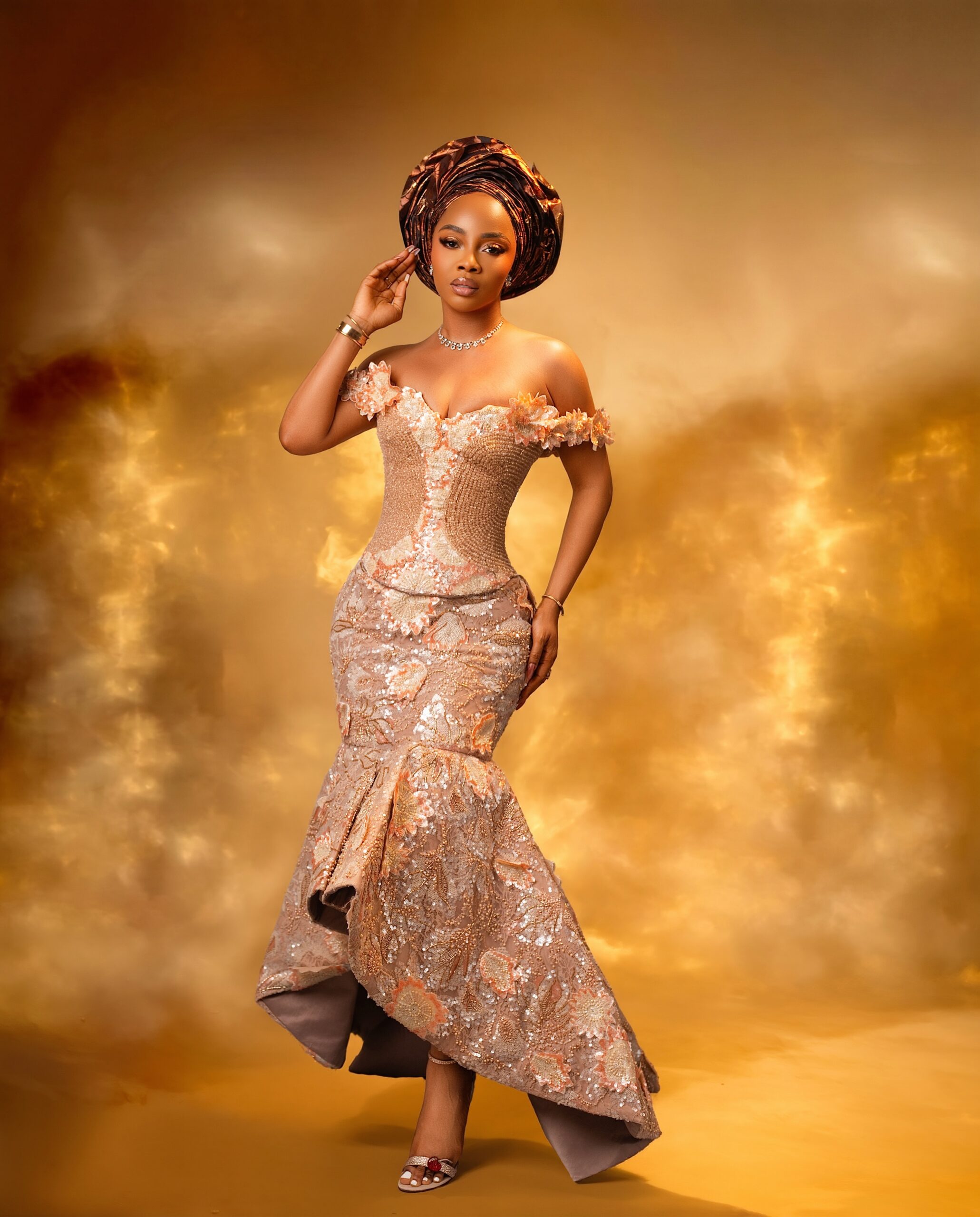 Toke Makinwa posing in an off-the-shoulder peach gown featuring structured gold-toned bodice panels and floral appliqués, styled with a bronze pleated gele.