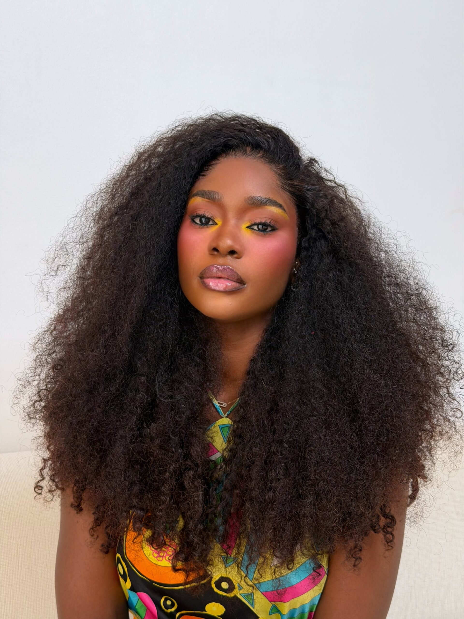 Dede Ashiogwu wears yellow and orange eyeshadow, rose blush, and a dark lined lip gloss with voluminous curly hair.