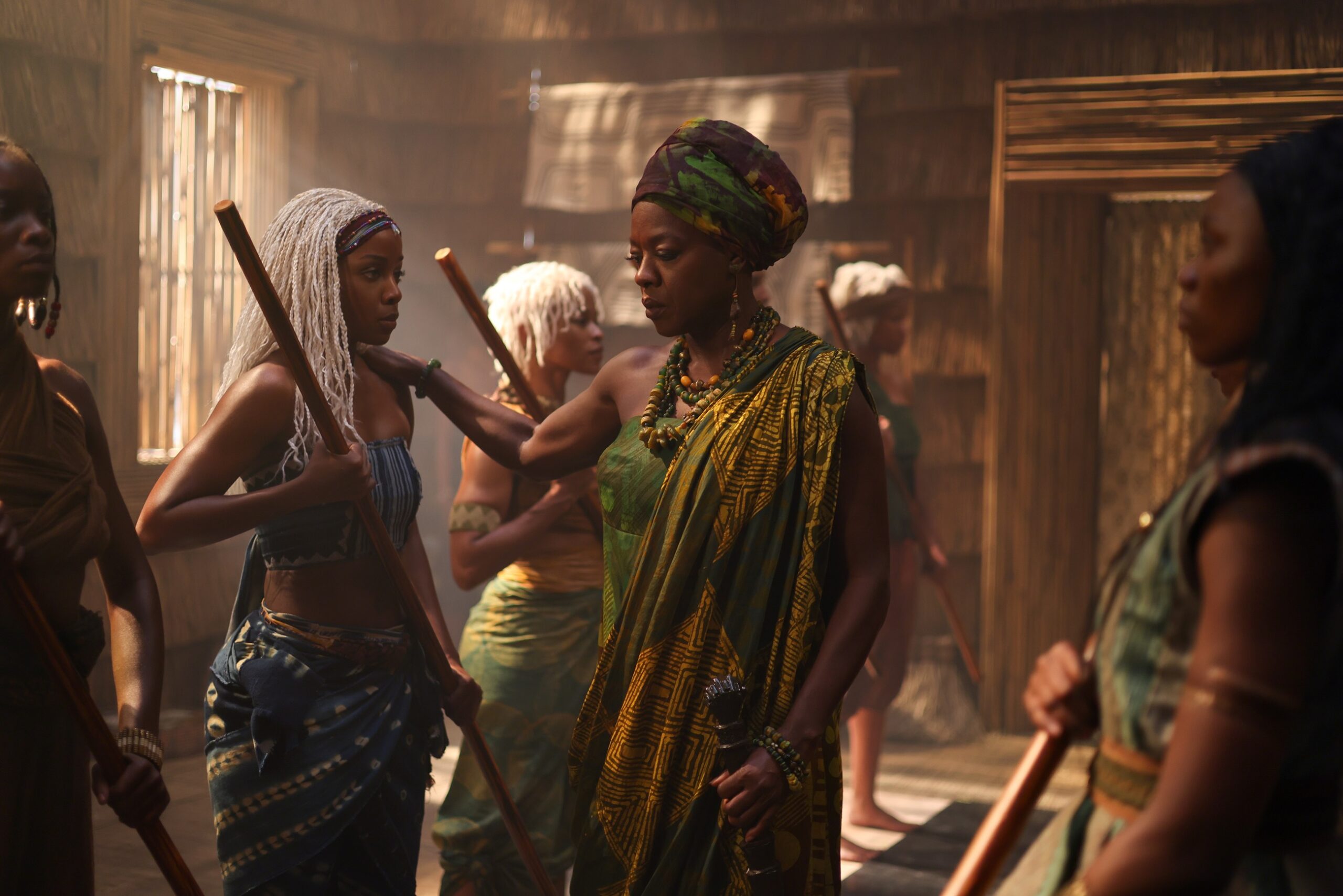 Viola Davis as Mama Agba in a green and yellow patterned wrap touching the shoulder of Thuso Mbedu as Zélie Adebola inside a wooden training pavilion.