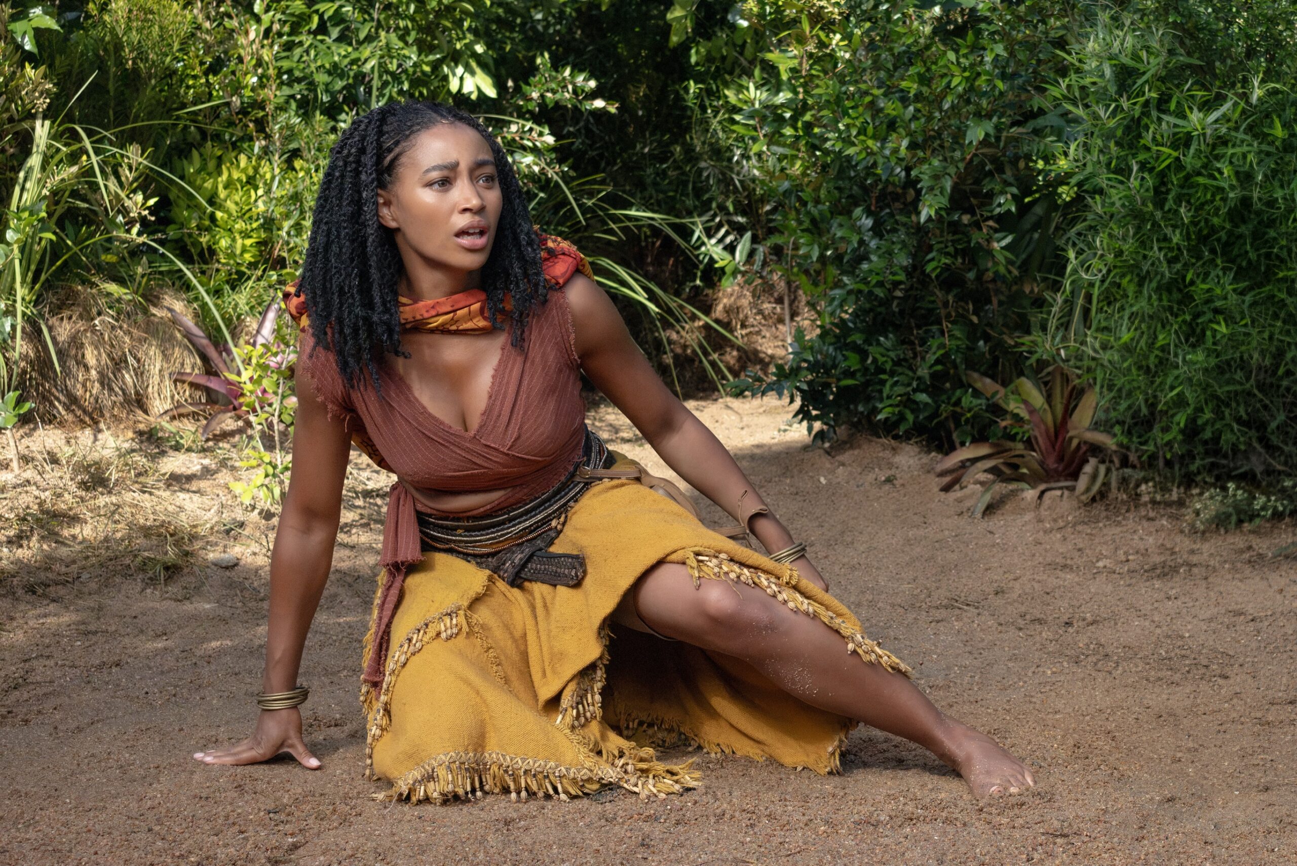 Amandla Stenberg as Princess Amari wearing a brown wrap top and yellow fringed skirt on a sandy path looking up with an intense expression.