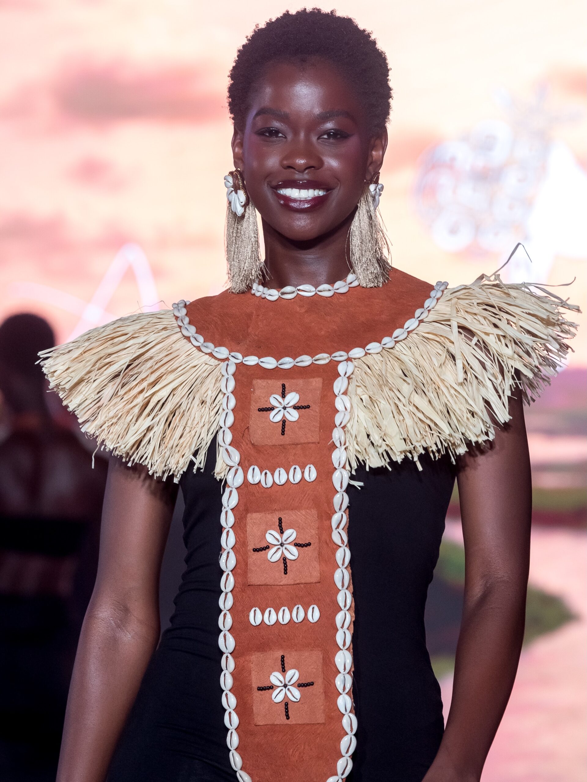 Kimberly Jael Aremo Acio wearing a terracotta suede bodice adorned with raffia fringe trim, cowrie shell embroidery, cowrie necklace, and gold tassel earrings during the Miss Universe Uganda 2026 traditional wear showcase.