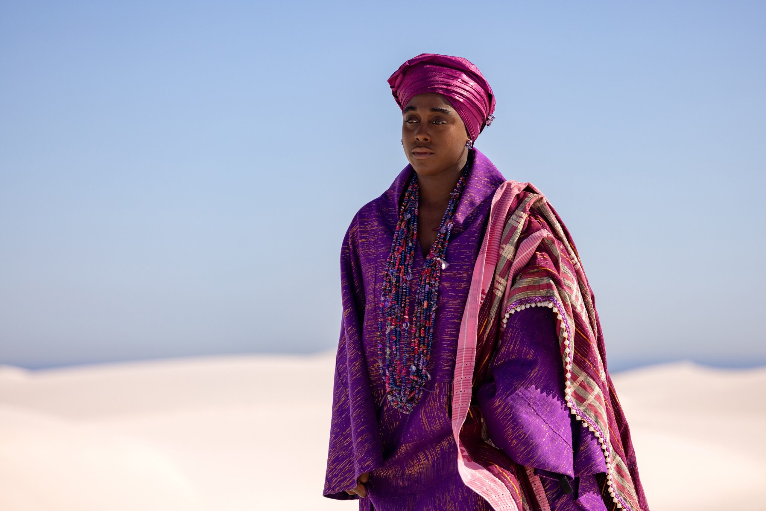 Lashana Lynch as Jumoke wearing a vibrant purple and pink traditional West African-inspired robe and headwrap in a white sand desert.