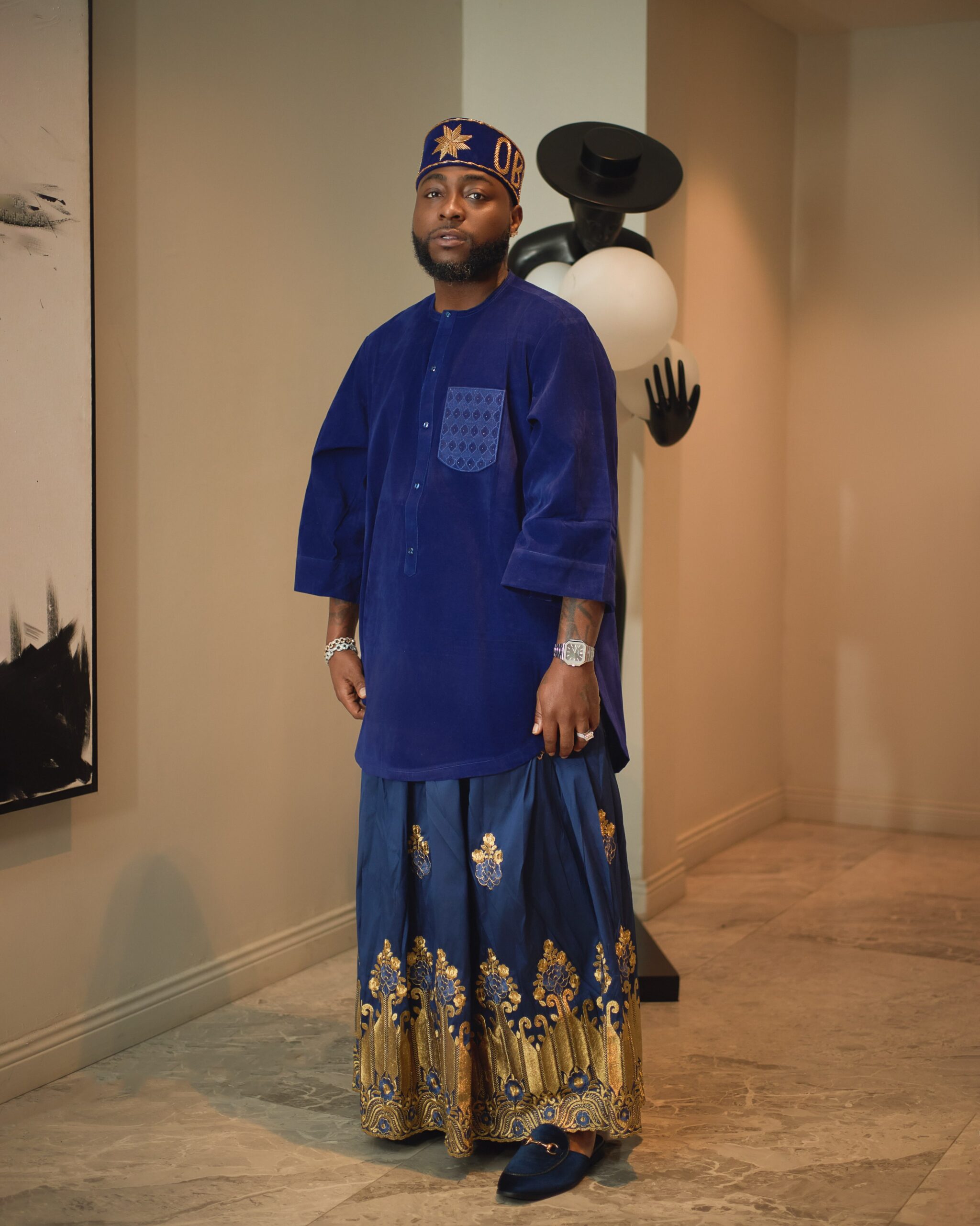 Davido showcasing a royal blue velvet three-quarter sleeve tunic with a geometric stitched chest pocket, silver wrist watch, and gold OBO cap.