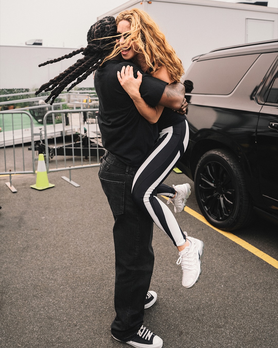 Burna Boy lifts Shakira off the ground in a celebratory hug next to a black SUV backstage at the FIFA World Cup stadium.