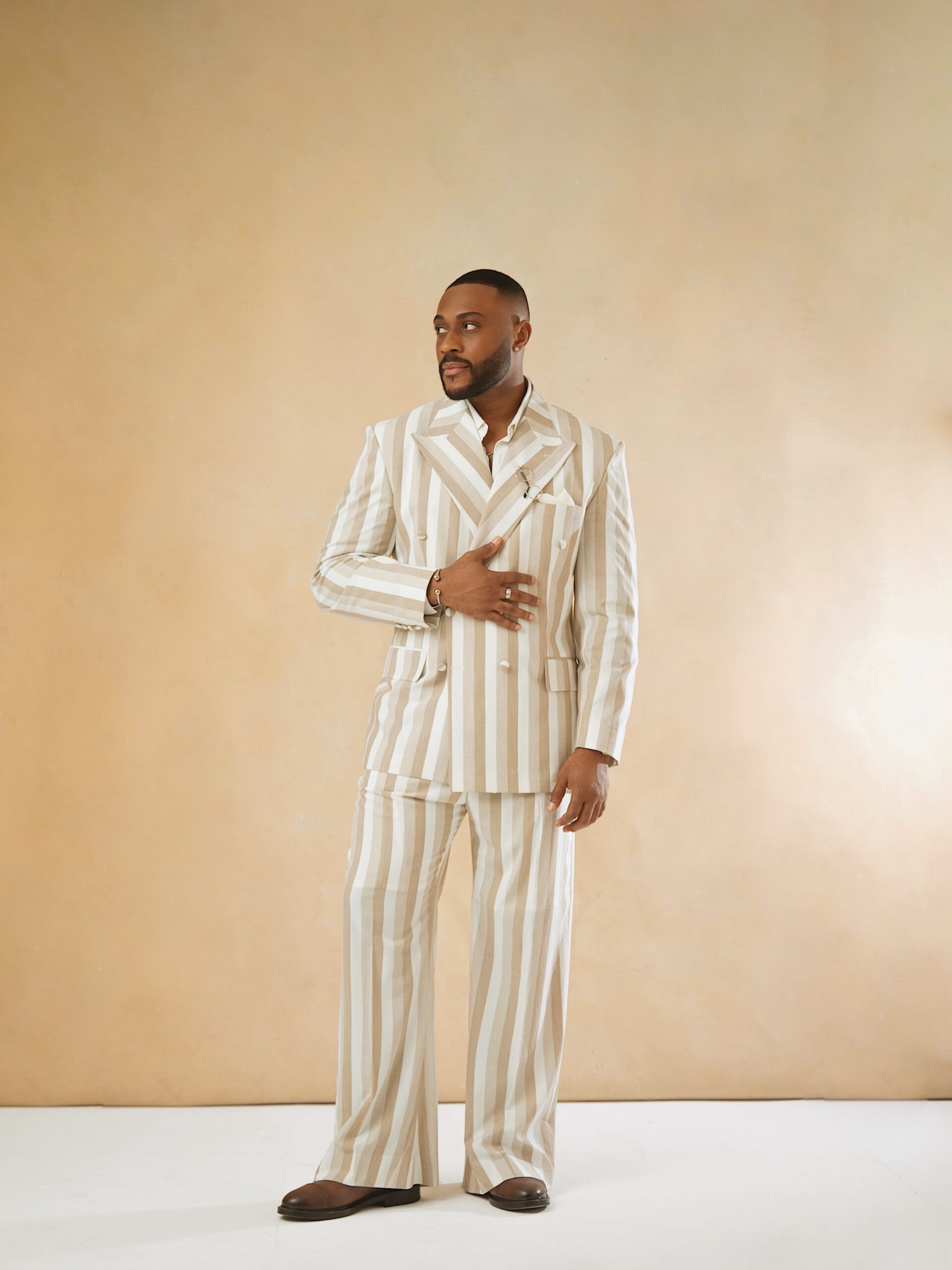 Kola Omotosho wearing a cream and beige vertical striped double-breasted suit by Oseyi Adams with wide-leg trousers and brown leather shoes.