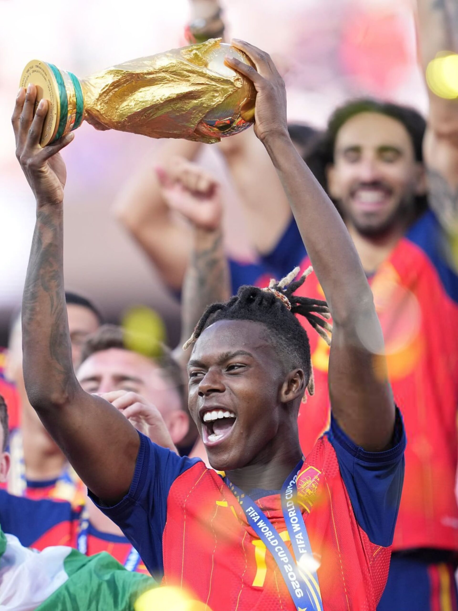 Nico Williams holds the gold FIFA World Cup trophy overhead surrounded by celebrating Spanish teammates.