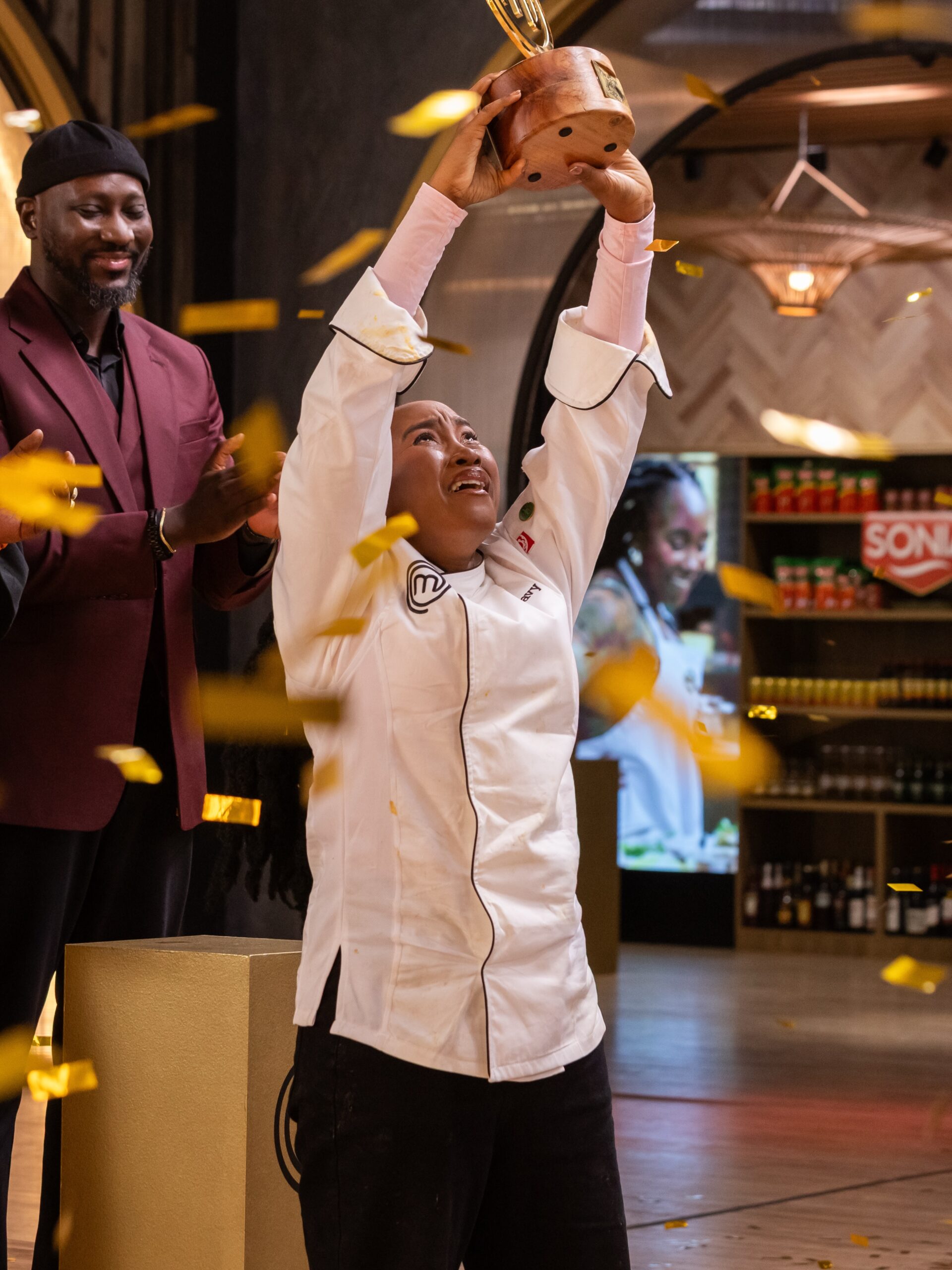 MasterChef Nigeria champion Favy Bessan holding up the trophy under falling confetti as Chef Eros applauds.