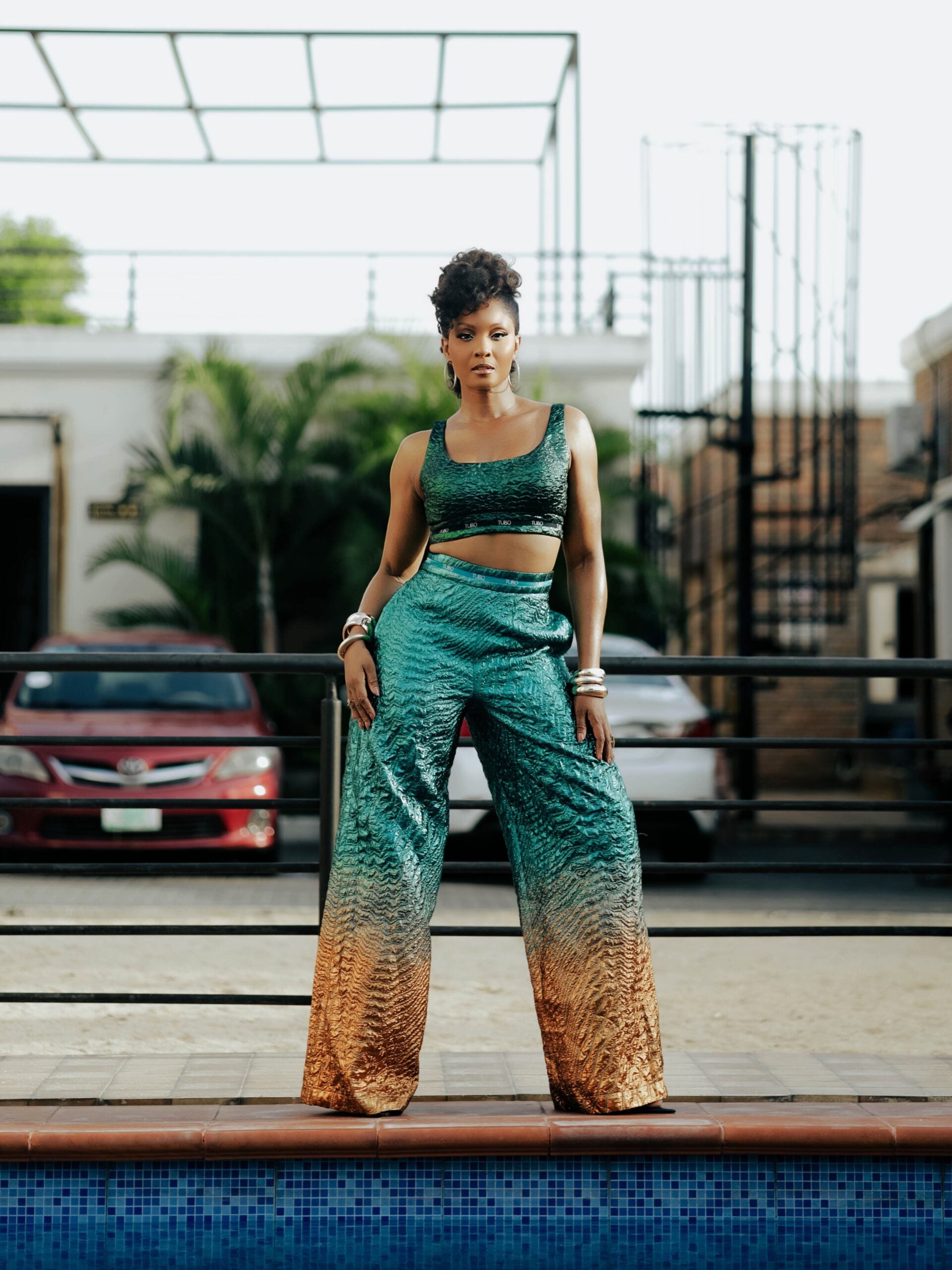 Osas Ighodaro standing beside a pool wearing a Tubo RTW two-piece set featuring a green metallic crop top and wide-leg trousers transitioning into bronze-gold ombré at the hem.