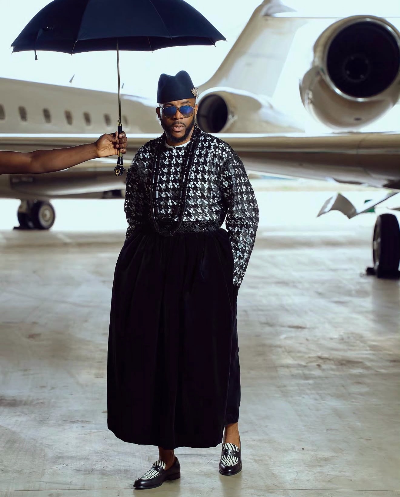 Ebuka Obi-Uchendu in a long-sleeved lattice sequin tunic and floor-length black velvet wrapper skirt paired with zebra-print leather loafers inside an aviation hangar for Big Brother Naija Season 11.