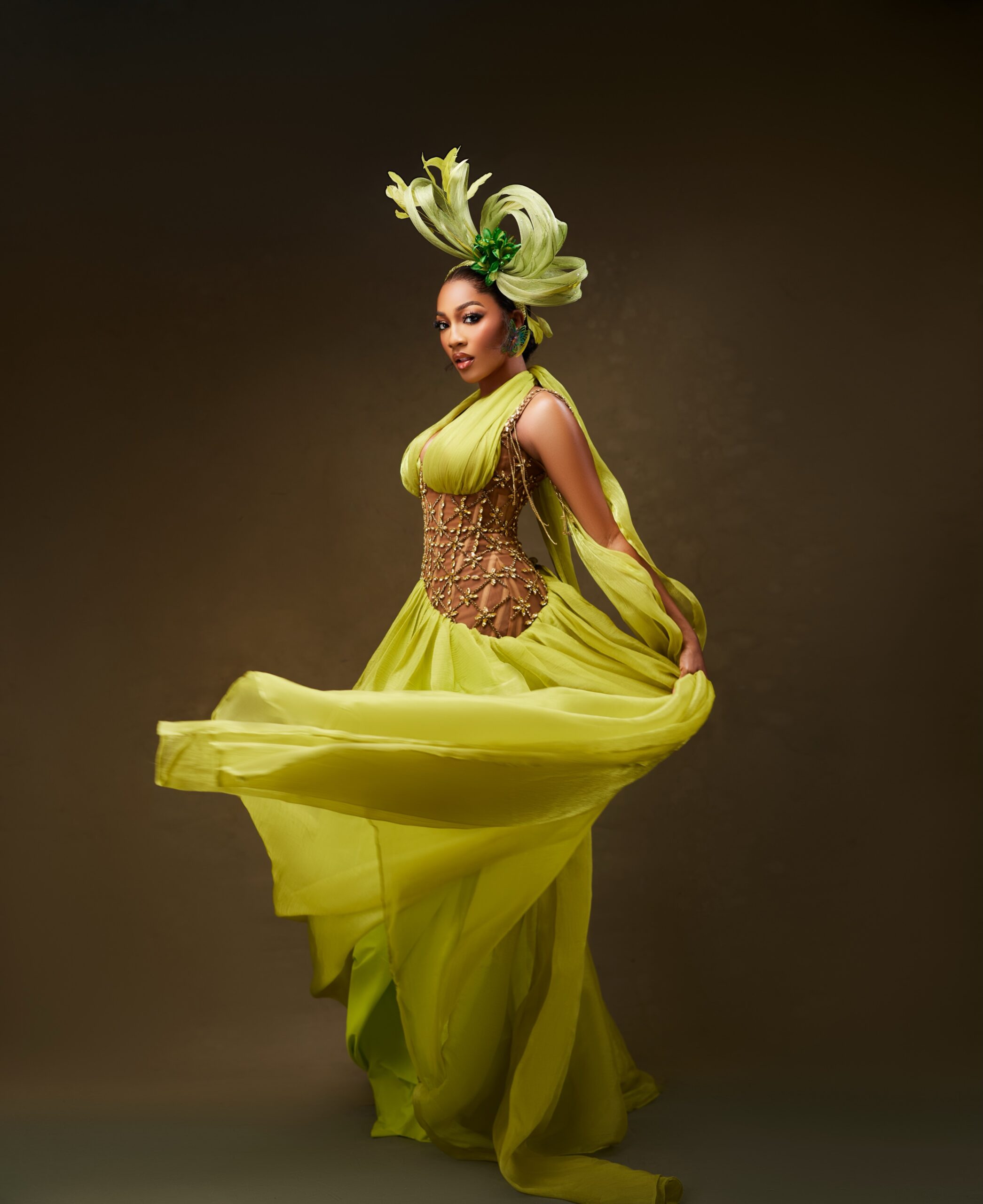 Hawa Magaji swirling a floor-length chartreuse yellow chiffon gown with an embellished lattice corset and a large green leaf fascinator.