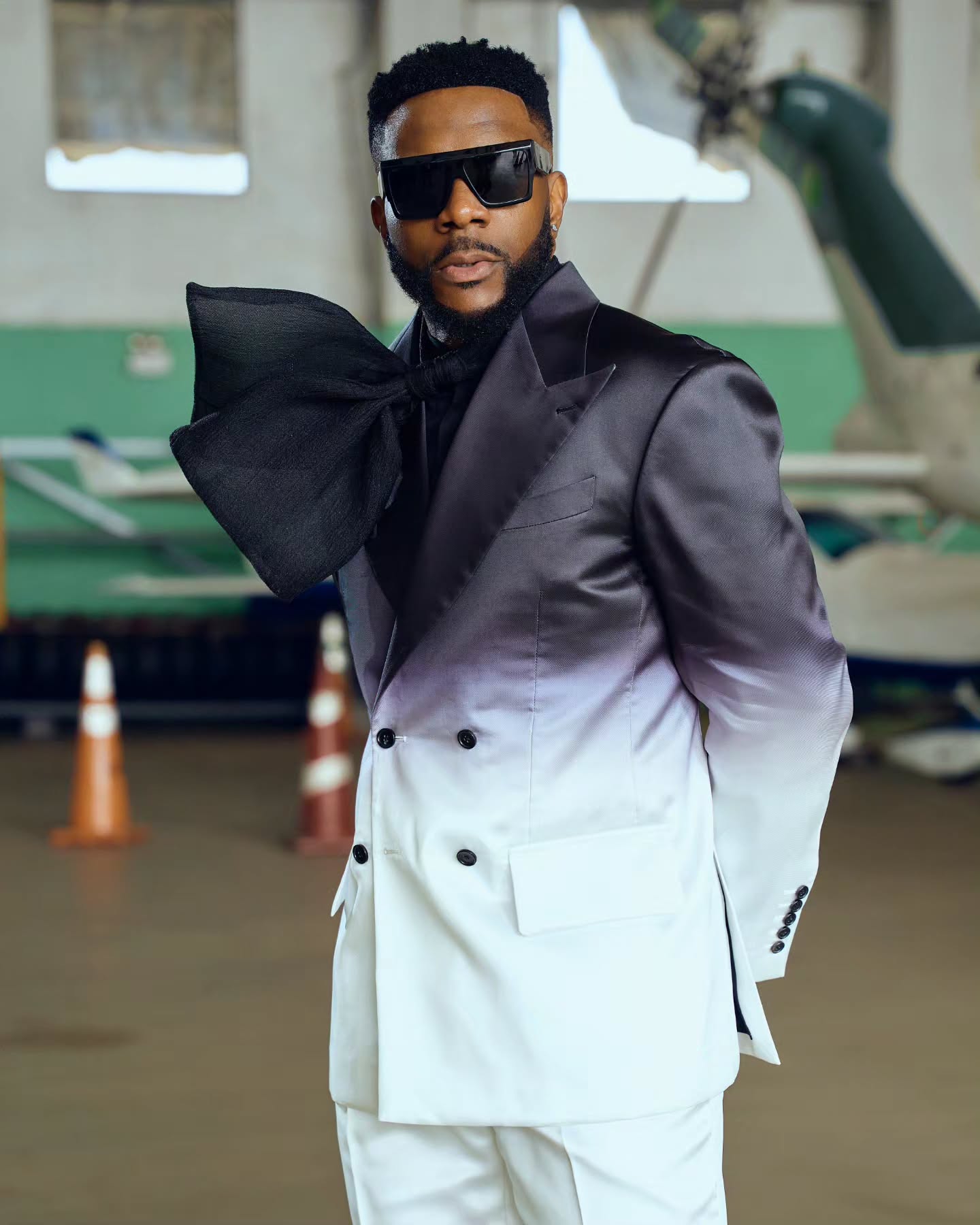 Ebuka Obi-Uchendu in an ATAFO ombré suit jacket with a statement fabric bow, square black sunglasses, and diamond ear studs posed in an airfield hangar for Big Brother Naija Season 11.