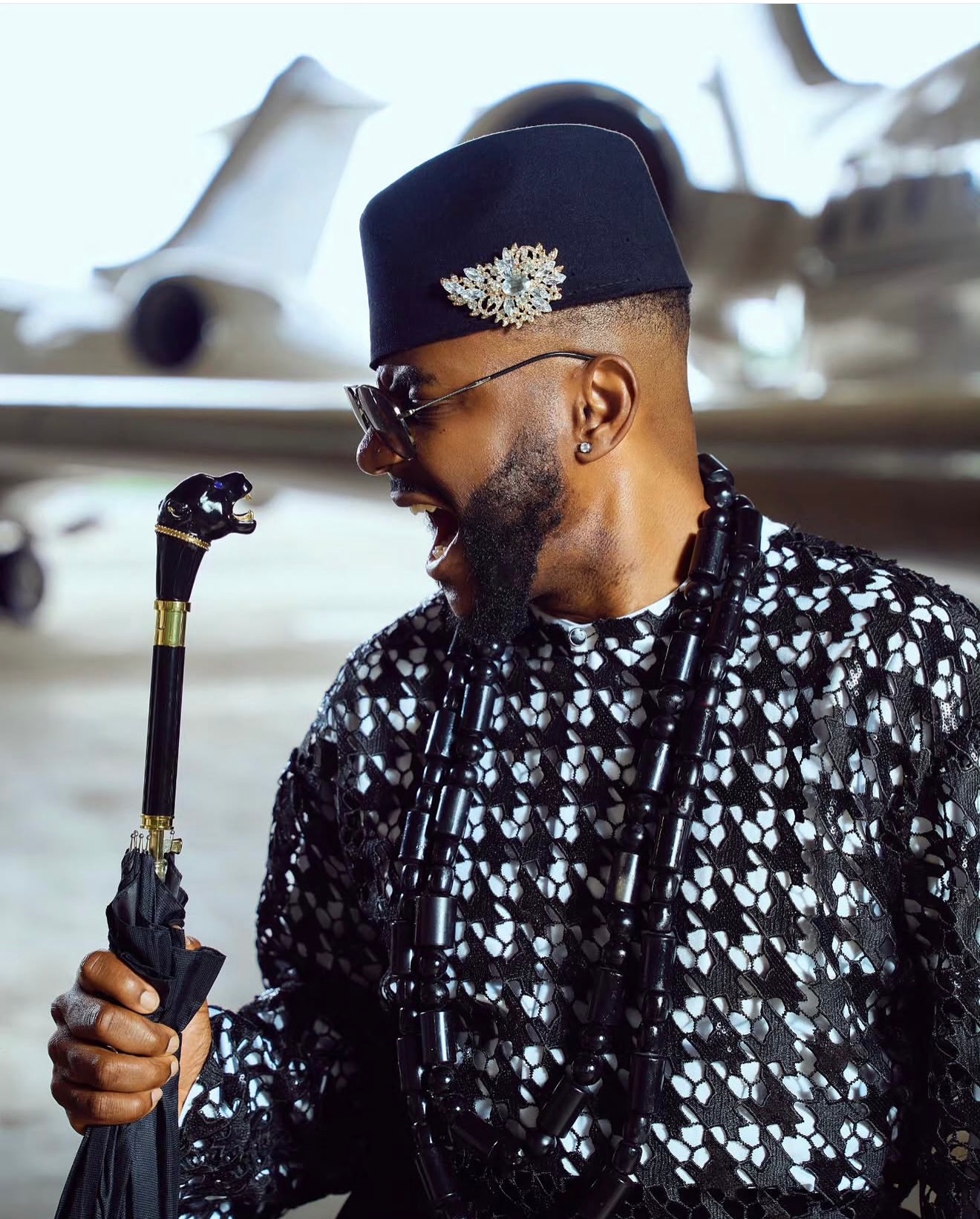 Ebuka Obi-Uchendu holding an umbrella with a sculpted animal handle, wearing a black traditional cap with a sparkling jewelled brooch and layered black beads for the Big Brother Naija Season 11 premiere.