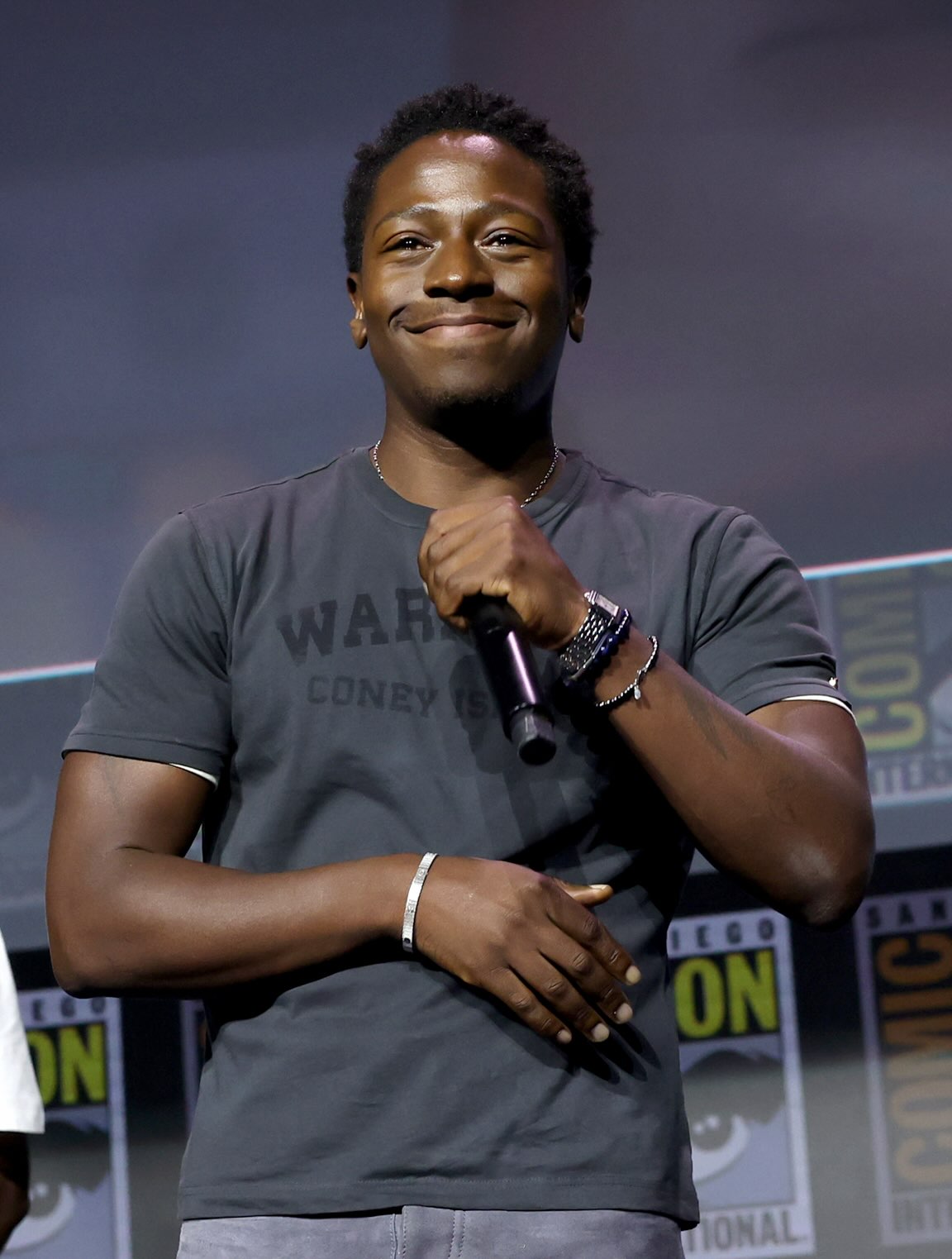 David Jonsson holding a microphone and smiling on stage in a dark grey graphic t-shirt and silver bracelet during the Black Panther 3 announcement at San Diego Comic-Con 2026.