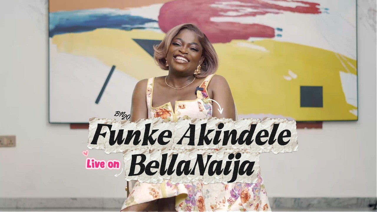 Funke Akindele smiling during her exclusive BellaNaija interview, wearing a floral mini dress and gold jewellery, seated against a colourful abstract painting backdrop