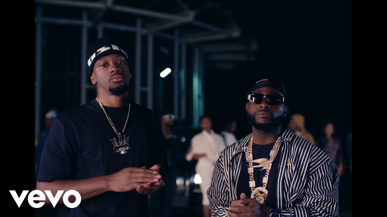 Houston-born singer NO11 in a black tee and beanie standing next to Davido who is wearing a striped shirt, sunglasses, and layered gold chains in the Gimme Dat Ting music video.