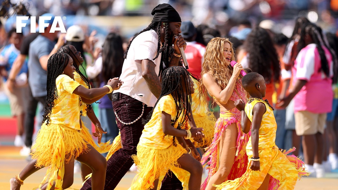 Shakira in a pink and yellow fringed outfit sings into a pink microphone alongside Burna Boy and Uganda's Ghetto Kids dancers on the pitch.